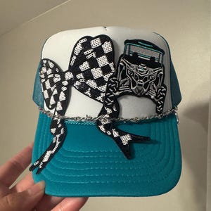 May include: A teal and white trucker hat with a black and white checkered bow and a black and silver off-road vehicle patch. The hat has a barbed wire detail across the front.