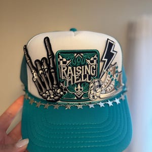 May include: Teal and white trucker hat with embroidered patches. Features a skeleton hand, lightning bolt, and the text "RAISING HELL". Silver star trim along the brim. A stylish accessory.