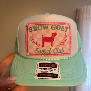 May include: A mint green and white trucker hat with the text "SHOW GOAT Social Club" on the front. The hat features a red goat graphic and a pink and white checkered design. The hat has a foam front and a snap closure.