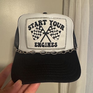 May include: A black and white trucker hat with a mesh back. The front panel features the text "START YOUR ENGINES" above crossed checkered flags, framed with silver glitter. A silver chain is attached to the brim.