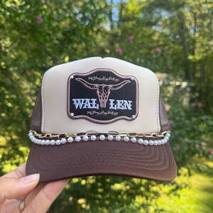 May include: A brown and tan trucker hat with a mesh back. The hat features a decorative patch with a steer skull and the name "WAL LEN". A gold chain and pearl trim adorns the brim.