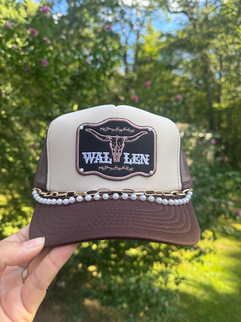 May include: A brown and tan trucker hat with a mesh back. The hat features a decorative patch with a steer skull and the name "WAL LEN". A gold chain and pearl trim adorns the brim.