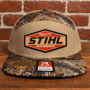 May include: A flat bill snapback hat with a camouflage pattern on the bill and top. The hat features a tan front panel with the "STIHL" logo in a red and orange hexagon. The hat is made by Richardson.