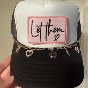 May include: A black and white trucker hat featuring a rectangular pink patch with the black embroidered text Let them and a small heart. A gold-toned chain is attached across the front, adorned with charms including a bow, a pearl heart, a smiley face, a lightning bolt, and a checkered heart.