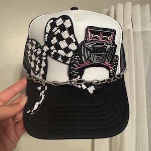 May include: A black and white trucker hat with a checkered flag bow and a sequined off-road vehicle design. The hat features a black brim, white front panel, and black mesh back. A silver chain is attached to the bow.
