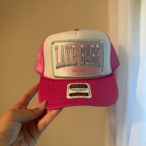 May include: A pink trucker hat with a white front panel. The hat features the text "LAKE BABE" in a teal, outlined font on a rectangular patch. The brim and mesh back are pink. A small tag is visible on the brim.