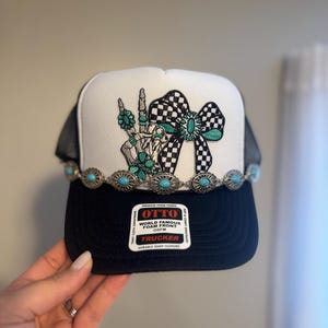 May include: Black and white trucker hat with a skeleton hand making a peace sign, a checkered bow, and turquoise accents. The hat has a decorative band with turquoise stones and the text "OTTO WORLD FAMOUS FOAM FRONT OSFM TRUCKER".