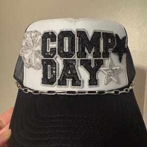 May include: Black and white trucker hat with the text "COMP DAY" in sequined letters. The hat features a silver chain, a star, and a decorative element on the side. The brim is black, and the top is white.