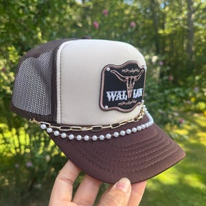 May include: A brown and tan trucker hat with a mesh back and a curved brim. The hat features a patch with a steer skull and the word "WAL LEN". A gold chain and pearl embellishment adorns the brim.