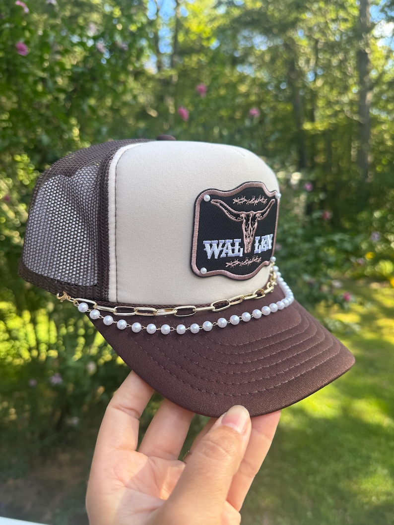 May include: A brown and tan trucker hat with a mesh back and a curved brim. The hat features a patch with a steer skull and the word "WAL LEN". A gold chain and pearl embellishment adorns the brim.