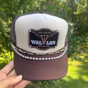May include: Brown and tan trucker hat with a mesh back and a curved brim. The hat features a patch with a steer skull and the text "WAL LEN". A gold chain and pearl beads adorn the brim.