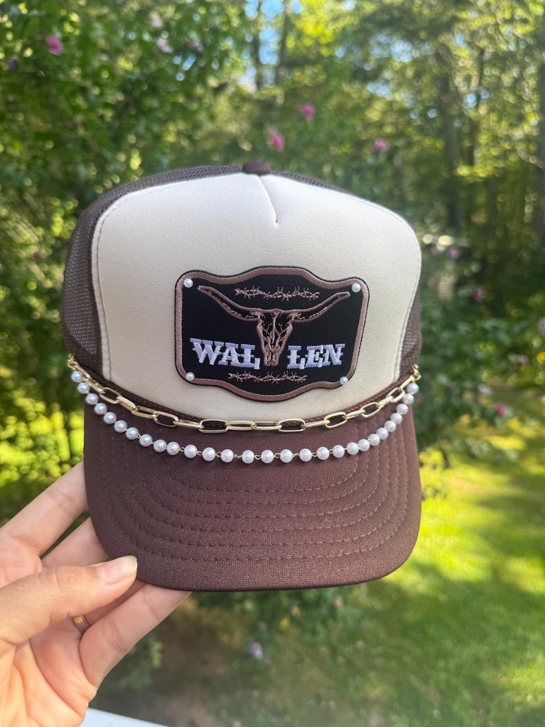 May include: Brown and tan trucker hat with a mesh back and a curved brim. The hat features a patch with a steer skull and the text "WAL LEN". A gold chain and pearl beads adorn the brim.