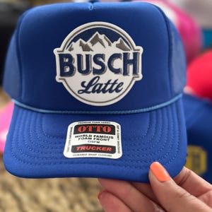May include: A blue trucker hat with the text "BUSCH Latte" in a white and gray circular emblem. The hat has a foam front and a snap closure. The hat is OSFM (One Size Fits Most).