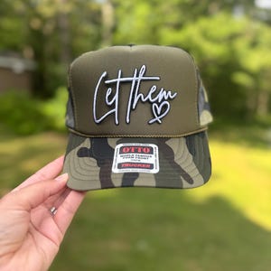 May include: Olive green and camouflage trucker hat with the text "Let them" in white script and a small heart. The hat has a mesh back and a curved brim. The brand tag is visible.
