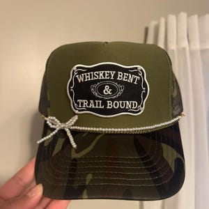 May include: A camouflage trucker hat with a black mesh back and a curved brim. The front features a patch with the text "WHISKEY BENT & TRAIL BOUND" in white lettering. A string of pearls and a bow accent the brim.
