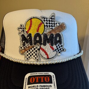 May include: A black and white trucker hat with a white front panel. The hat features a beaded design with the word "MAMA", a softball, baseball, and a bat. The brim is lined with pearls. The hat has the text "OTTO TRUCKER".