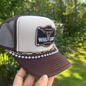 May include: A brown and tan trucker hat with a mesh back and a curved brim. The hat features a patch with a bull skull design and the text "WAL LEN". A gold chain and pearl bead chain are attached to the brim.
