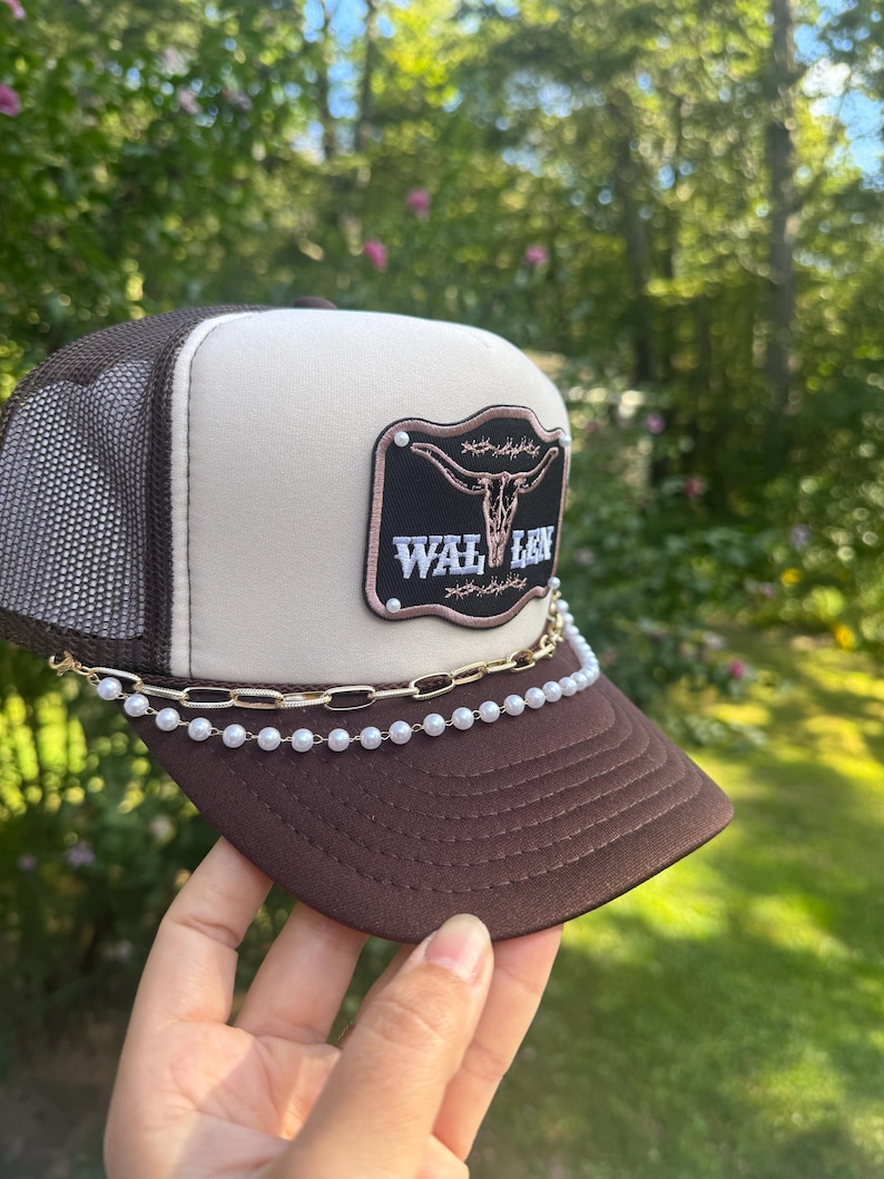 May include: A brown and tan trucker hat with a mesh back and a curved brim. The hat features a patch with a bull skull design and the text "WAL LEN". A gold chain and pearl bead chain are attached to the brim.