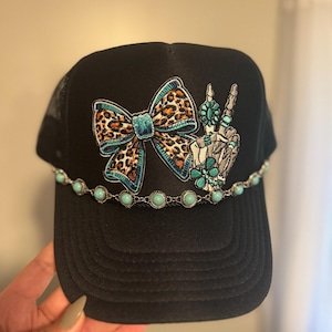 May include: Black trucker hat with a leopard print bow and a skeleton hand making a peace sign. The hat features turquoise accents and a chain of turquoise beads along the brim. The hat is a stylish accessory.
