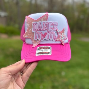 May include: A pink and white trucker hat with the words "DANCE MOM" in pink letters. The hat has a pink brim and a white mesh back. Two pink star-shaped embellishments are on the front.