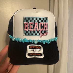 May include: A black and white trucker hat with the word "BEACH" in pink embroidery. The hat has a turquoise stone trim and a black mesh back. The hat has a "Durable Snap Closure" and "World Famous Foam Front".