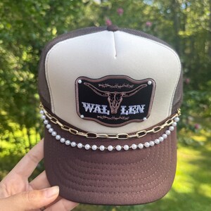 May include: Brown trucker hat with a white front panel and a mesh back. The hat features a decorative patch with a bull skull and the text "WAL LEN". A gold chain and pearl beads adorn the brim.