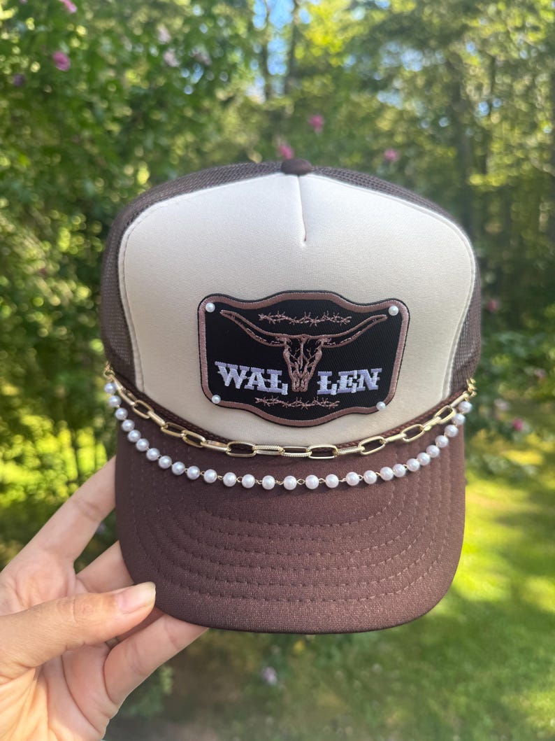 May include: Brown trucker hat with a white front panel and a mesh back. The hat features a decorative patch with a bull skull and the text "WAL LEN". A gold chain and pearl beads adorn the brim.