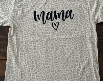 May include: A light-colored t-shirt with a leopard print pattern. The word "mama" is printed in black script with a small heart underneath. The shirt has a crew neck and short sleeves.
