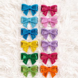 May include: A collection of colorful sequined hair bows in various shades, including blue, pink, yellow, green, and orange. The bows are arranged on a fluffy white surface, showcasing their vibrant colors and textures. These accessories are perfect for adding a touch of sparkle.