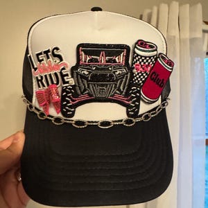 May include: Black and white trucker hat with a chain detail. The hat features patches with the text "Lets Ride", a pink and black off-road vehicle, and two red and black "530 Club" beverage cans. The hat has a black brim and white mesh back.