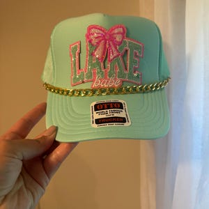 May include: A mint green trucker hat with the text "LAKE babe" in pink and sequined letters, topped with a pink sequined bow. A gold chain is attached to the brim. The hat has a snap closure and a tag that reads "OTTO".