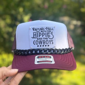 May include: A burgundy and white trucker hat with a black chain detail. The hat features the text "Raising Hell with the Hippies and the Cowboys" on a patch. The hat is held by a hand against a blurred green background.