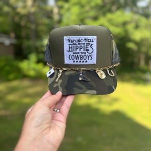 May include: A camouflage trucker hat with a black mesh back and a square patch that reads "Raising Hell with the Hippies and the Cowboys." The hat is adorned with a gold chain and charms, including a key, a boot, and a horseshoe.