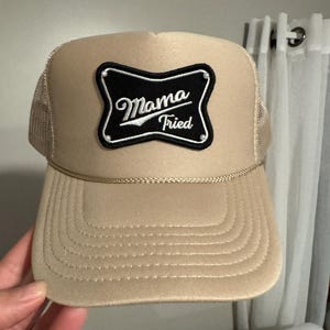 May include: Tan trucker hat with a mesh back and a curved brim. The front features a black and white patch with the words "Mama Tried" in a script font. A gold-colored rope detail accents the brim.