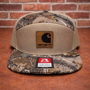 May include: A Carhartt flat bill snapback hat with a camouflage pattern on the bill and top. The front panel is tan with a brown leather patch featuring the Carhartt logo. The hat has a mesh back and a Richardson tag.