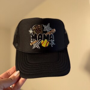 May include: Black trucker hat with a mesh back and a curved brim. The front features the word "MAMA" in sequins, along with a baseball bat, a softball, and checkered flag designs. The hat is held by a hand.