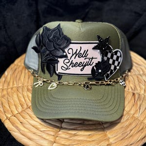 May include: A green trucker hat with a black rose, a pink patch that says "Well Sheeyit", a black and white checkered heart, and a gold chain with charms.
