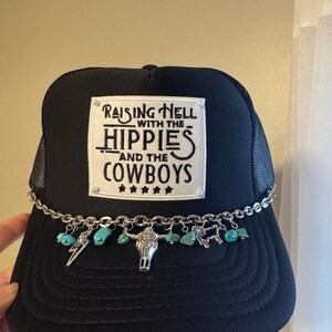 May include: Black trucker hat with a mesh back and a decorative chain. The hat features a patch with the text "Raising Hell with the Hippies and the Cowboys." Turquoise stones and silver charms, including a bull skull, adorn the brim.