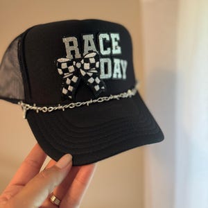 May include: Black trucker hat with the text "RACE DAY" in silver glitter. A checkered flag bow is centered below the text. A silver chain is attached to the brim of the hat. The hat is held by a hand.