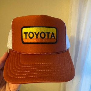 May include: A burnt orange trucker hat with a white mesh back. The front panel features a rectangular patch with the word "TOYOTA" in bold, black letters. The patch has a yellow and orange gradient background.