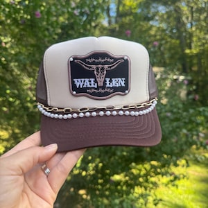 May include: A brown and tan trucker hat with a decorative pearl chain and the word "WAL LEN" on a black patch with a steer skull design. The hat has a mesh back and is being held up by a hand. Morgan Wallen Trucker Hat with Chain: Wallen trucker hat