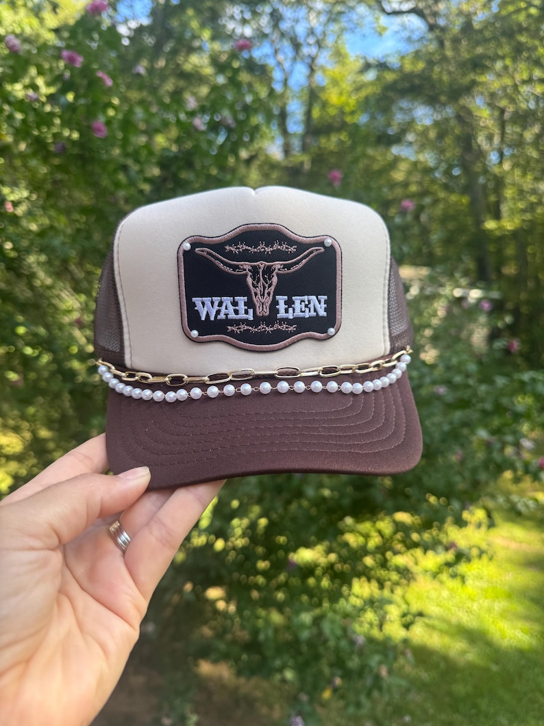 May include: A brown and tan trucker hat with a decorative pearl chain and the word "WAL LEN" on a black patch with a steer skull design. The hat has a mesh back and is being held up by a hand. Morgan Wallen Trucker Hat with Chain: Wallen trucker hat