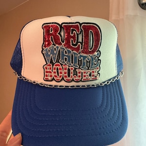 May include: A blue and white trucker hat with the words "RED WHITE BOOJEE" in red, white, and blue sequined letters. The hat has a blue brim and a silver chain detail.