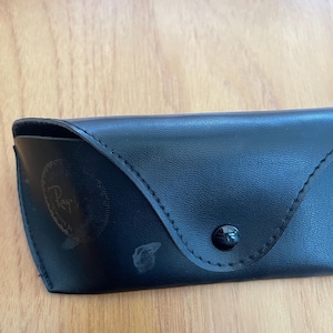 Puede incluir: Estuche de gafas de sol de cuero negro con cierre de bot&oacute;n a presi&oacute;n. El estuche tiene un borde cosido y un logotipo descolorido en la parte delantera. Dise&ntilde;ado para proteger gafas.