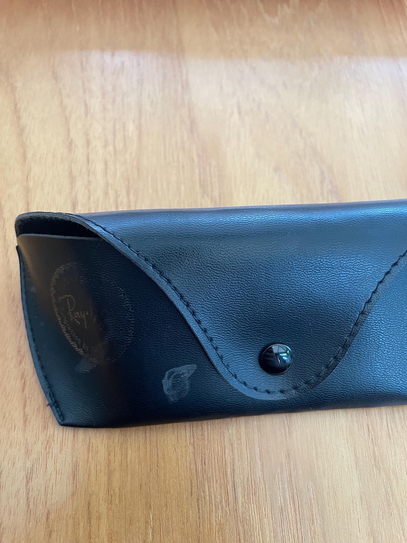 Puede incluir: Estuche de gafas de sol de cuero negro con cierre de bot&oacute;n a presi&oacute;n. El estuche tiene un borde cosido y un logotipo descolorido en la parte delantera. Dise&ntilde;ado para proteger gafas.