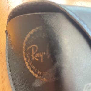 Puede incluir: Primer plano de un estuche negro para gafas con el logotipo dorado de Ray-Ban. El estuche est&aacute; hecho de un material liso, posiblemente cuero. El logotipo es circular con el nombre de la marca en una fuente estilizada.
