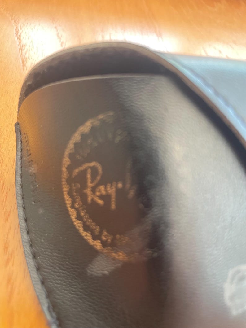 Puede incluir: Primer plano de un estuche negro para gafas con el logotipo dorado de Ray-Ban. El estuche est&aacute; hecho de un material liso, posiblemente cuero. El logotipo es circular con el nombre de la marca en una fuente estilizada.