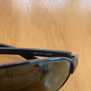 Puede incluir: Primer plano de gafas de sol negras con lentes oscuras. El texto "RB 3101 W3060" est&aacute; impreso en la montura. Las gafas de sol est&aacute;n sobre una superficie de madera.
