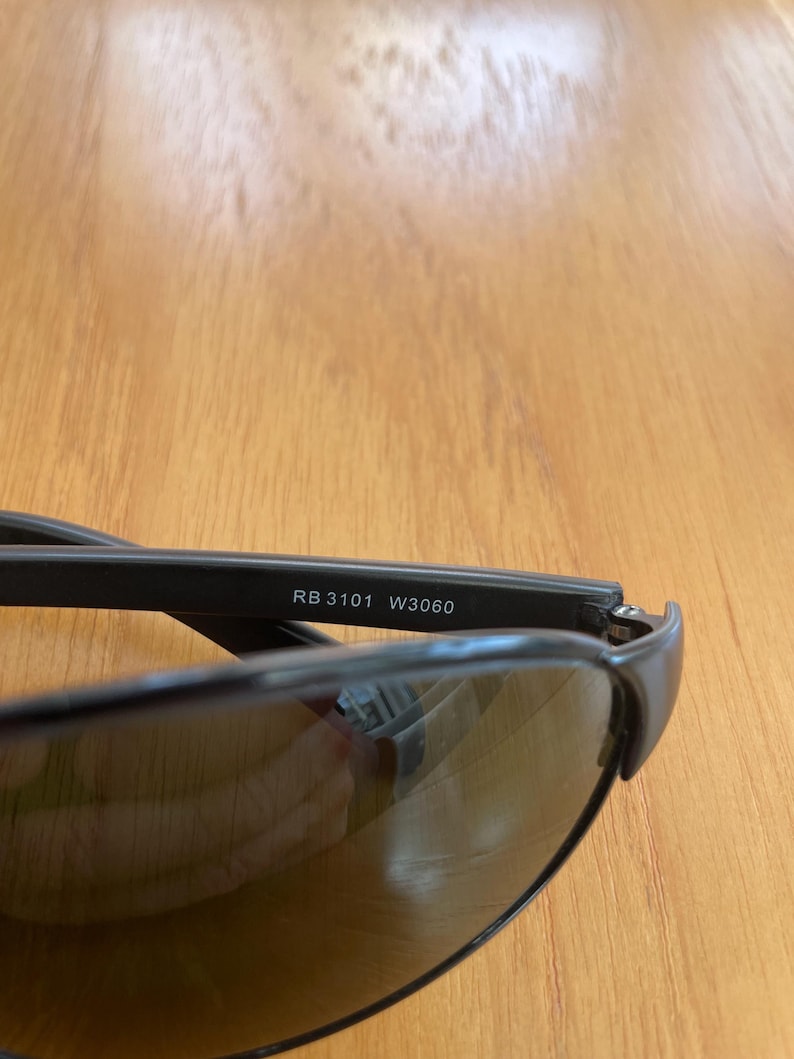 Puede incluir: Primer plano de gafas de sol negras con lentes oscuras. El texto "RB 3101 W3060" est&aacute; impreso en la montura. Las gafas de sol est&aacute;n sobre una superficie de madera.