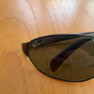 Puede incluir: Primer plano de gafas de sol Ray-Ban con lentes oscuras y montura negra. El logotipo de Ray-Ban es visible en la lente izquierda. Las gafas de sol est&aacute;n sobre una superficie de madera.